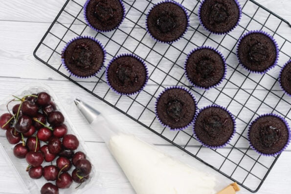 chocolate cupcakes on a wire rack with cherries on the side and a piping bag full of whipped cream.