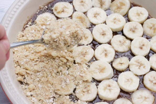 adding crumble to the top of bananas arranged in a serving dish.